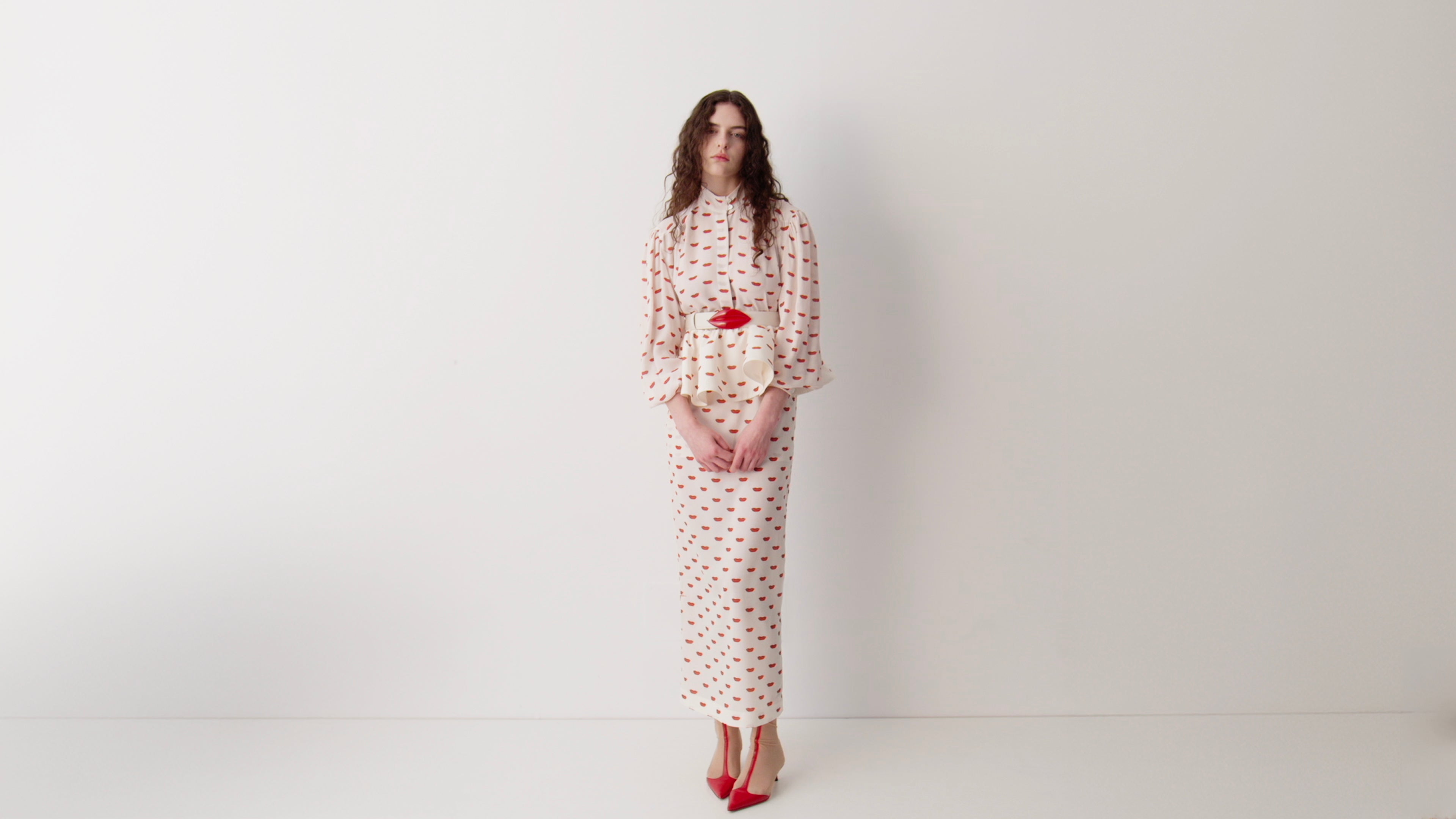 Model wearing a cream maxi dress with red lip print, belted at the waist, styled with red boots.